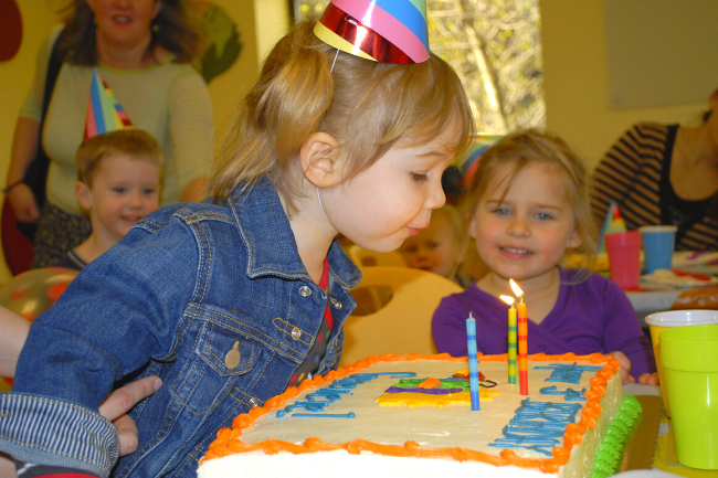 A child with blonde pigtails, wearing a blue jean jacket and a striped party hat leans over a cake to blow out three candles.