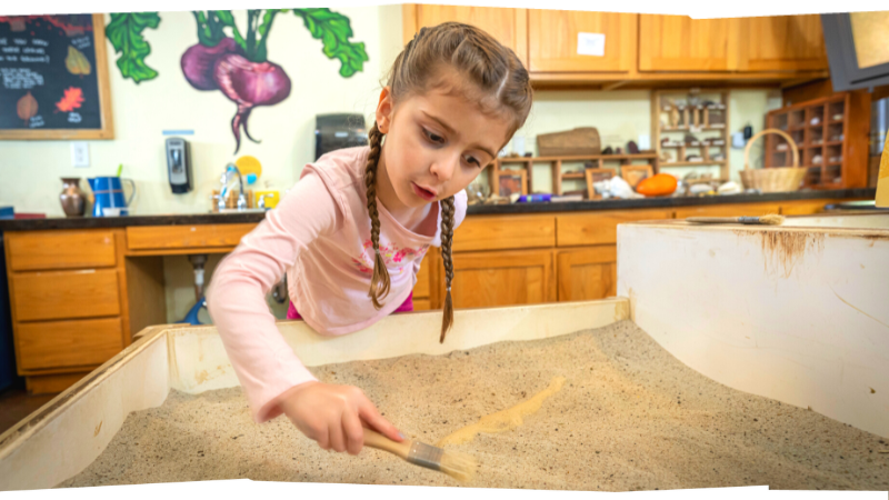 A little girl leans over the fossil hunting sand table, a brush in her hand.